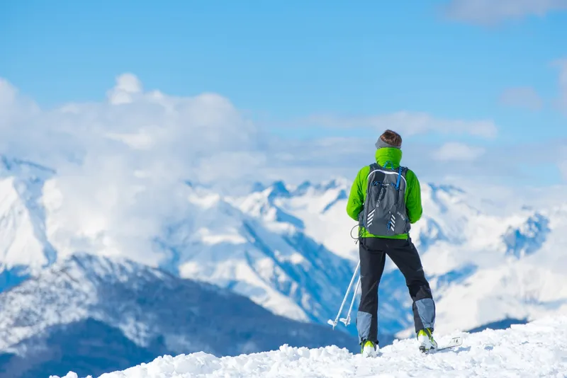 Person steht in Winterkleidung auf schneebedecktem Berg, blickt auf verschneite Gipfel.