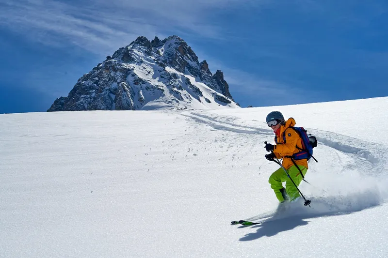 Person fährt Ski im Schnee neben verschneitem Berggipfel.