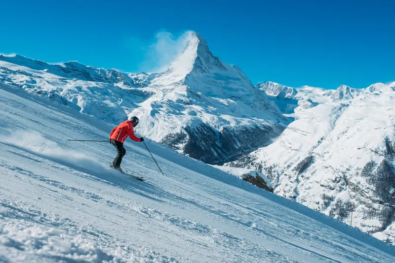 Person in roter Jacke beim Skifahren vor schneebedecktem Bergpanorama.